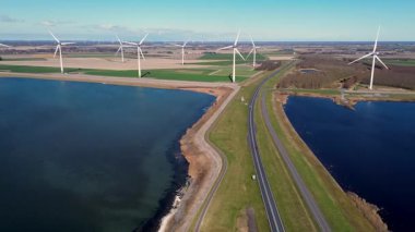 Yeşil tarlalarda rüzgar türbinleriyle kaplı iki su kütlesi arasında bir kıyı yolu var. Sahnede yenilenebilir enerji, altyapı ve doğa açık mavi gökyüzü altında vurgulanıyor..
