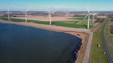 Yeşil tarlalarda rüzgar türbinleriyle kaplı iki su kütlesi arasında bir kıyı yolu var. Sahnede yenilenebilir enerji, altyapı ve doğa açık mavi gökyüzü altında vurgulanıyor..