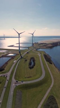 Yeşil tarlalarda rüzgar türbinleriyle kaplı iki su kütlesi arasında bir kıyı yolu var. Sahnede yenilenebilir enerji, altyapı ve doğa açık mavi gökyüzü altında vurgulanıyor..