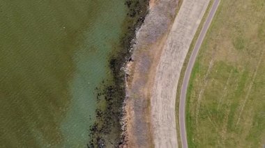 Yeşil suyu, kayalık plajı, kıvrımlı yolu ve çimenli arazisi olan bir kıyı şeridini gösteren bir hava görüntüsü. Yukarıdan aşağı perspektif, dokuları ve doğal desenleri vurgular.