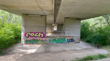 Grafiti kaplı sütunları olan beton bir köprü yeşil bir alanda duruyor. Güneş ışığı ağaçların arasından süzülüp yere gölge düşürür. Kentsel ve doğal elementlerin karışımı çarpıcı bir kontrast yaratır.