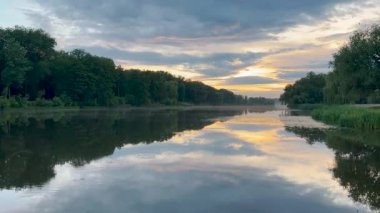 Sakin bir göl, yemyeşil ağaçlarla çevrili günbatımının altın tonlarını yansıtır. Suda hafif dalgalanmalar oluşur ve gökyüzü yumuşak maviler ve ılık portakalların karışımıdır. Huzurlu bir manzara yaratır.