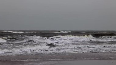 Dalgalar gri, bulutlu bir gökyüzünün altında kıyıya çarpar. Yüzeyde beyaz köpük oluşurken deniz dalgalı görünür. Ufuk uzaklıktan görülebiliyor, derinlik katıyor..
