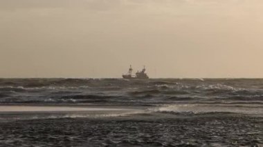 Bir balıkçı teknesi, sisli bir gökyüzü altında dalgalar arasında yelken açar. Rüzgâr türbinleri uzak arkaplanda sisten zar zor görülür. Deniz kuşları su üzerinde uçar ve deniz manzarasına eklenir.