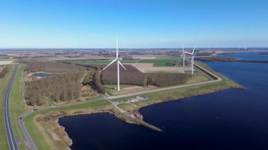 Yeşil tarlalarda rüzgar türbinleriyle kaplı iki su kütlesi arasında bir kıyı yolu var. Sahnede yenilenebilir enerji, altyapı ve doğa açık mavi gökyüzü altında vurgulanıyor..