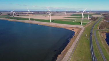 Yeşil tarlalarda rüzgar türbinleriyle kaplı iki su kütlesi arasında bir kıyı yolu var. Sahnede yenilenebilir enerji, altyapı ve doğa açık mavi gökyüzü altında vurgulanıyor..