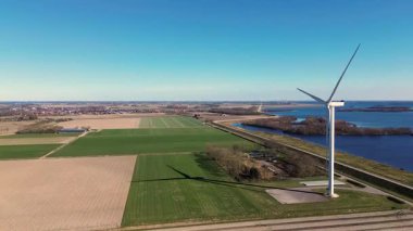Kırsal bir alanda, yeşil tarlaları, bir nehri ve uzak bir köyü olan büyük bir rüzgar türbininin hava görüntüleri. Yel değirmeni açık mavi bir gökyüzü altında dönerek, doğadaki sürdürülebilir enerjiyi vurgular..