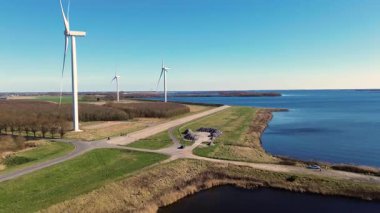 Yeşil tarlalarda rüzgar türbinleriyle kaplı iki su kütlesi arasında bir kıyı yolu var. Sahnede yenilenebilir enerji, altyapı ve doğa açık mavi gökyüzü altında vurgulanıyor..