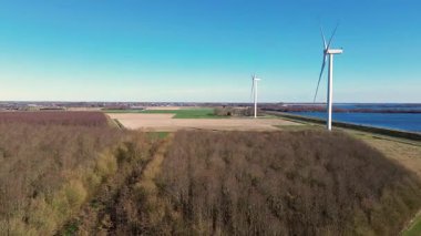 Bir dron, kırsal bir alanda rüzgar türbinlerinin çarpıcı bir hava görüntüsünü yakalar. Yeşil alanlar, bir nehir ve parlak mavi bir gökyüzü yenilenebilir enerji ve sürdürülebilirliği vurgulayan güzel bir manzara yaratır..