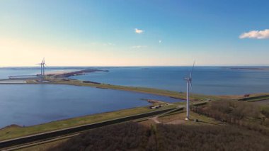Yeşil tarlalarda rüzgar türbinleriyle kaplı iki su kütlesi arasında bir kıyı yolu var. Sahnede yenilenebilir enerji, altyapı ve doğa açık mavi gökyüzü altında vurgulanıyor..