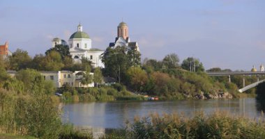 Bereketli bir nehir kenarı manzarası, bir köprü ve tarihi binalar, içinde kubbesi olan bir kilise de var. Sakin su güneşli günün sakin atmosferini yansıtır..