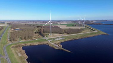 Rüzgar türbinlerinin tarlaların, yolların ve su yollarının üzerinde döndüğü çarpıcı bir kıyı manzarası. Açık mavi gökyüzü ve sakin sular resim gibi ve sürdürülebilir bir enerji sahnesi yaratır..