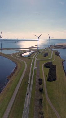 Yeşil tarlalarda rüzgar türbinleriyle kaplı iki su kütlesi arasında bir kıyı yolu var. Sahnede yenilenebilir enerji, altyapı ve doğa açık mavi gökyüzü altında vurgulanıyor..