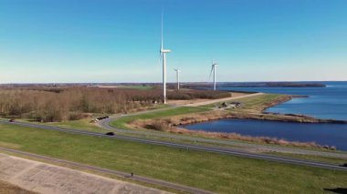 Rüzgar türbinlerinin tarlaların, yolların ve su yollarının üzerinde döndüğü çarpıcı bir kıyı manzarası. Açık mavi gökyüzü ve sakin sular resim gibi ve sürdürülebilir bir enerji sahnesi yaratır..