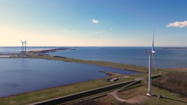 Yeşil tarlalarda rüzgar türbinleriyle kaplı iki su kütlesi arasında bir kıyı yolu var. Sahnede yenilenebilir enerji, altyapı ve doğa açık mavi gökyüzü altında vurgulanıyor..