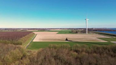 Kırsal bir alanda, yeşil tarlaları, bir nehri ve uzak bir köyü olan büyük bir rüzgar türbininin hava görüntüleri. Yel değirmeni açık mavi bir gökyüzü altında dönerek, doğadaki sürdürülebilir enerjiyi vurgular..