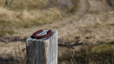 Paslı bir at nalı, kurumuş, kırsal bir arazide yıpranmış ahşap bir direğin üzerinde duruyor. Sıcak güneş ışığı yumuşak gölgeler bırakır, eski ahşap ve demirin dokusunu aydınlatır. Huzurlu bir kır manzarası..