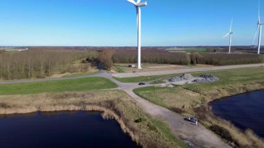 Yeşil tarlalarda rüzgar türbinleriyle kaplı iki su kütlesi arasında bir kıyı yolu var. Sahnede yenilenebilir enerji, altyapı ve doğa açık mavi gökyüzü altında vurgulanıyor..