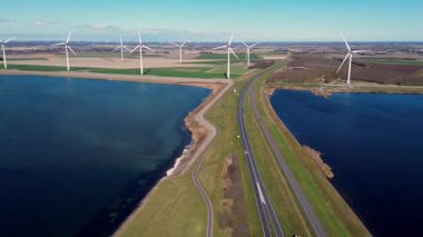 Yeşil tarlalarda rüzgar türbinleriyle kaplı iki su kütlesi arasında bir kıyı yolu var. Sahnede yenilenebilir enerji, altyapı ve doğa açık mavi gökyüzü altında vurgulanıyor..