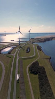 Yeşil tarlalarda rüzgar türbinleriyle kaplı iki su kütlesi arasında bir kıyı yolu var. Sahnede yenilenebilir enerji, altyapı ve doğa açık mavi gökyüzü altında vurgulanıyor..