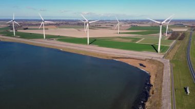 Yeşil tarlalarda rüzgar türbinleriyle kaplı iki su kütlesi arasında bir kıyı yolu var. Sahnede yenilenebilir enerji, altyapı ve doğa açık mavi gökyüzü altında vurgulanıyor..