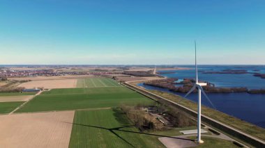 Kırsal bir alanda, yeşil tarlaları, bir nehri ve uzak bir köyü olan büyük bir rüzgar türbininin hava görüntüleri. Yel değirmeni açık mavi bir gökyüzü altında dönerek, doğadaki sürdürülebilir enerjiyi vurgular..