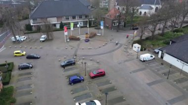 Park halindeki arabaların, bisikletçilerin bulunduğu sakin bir kasaba meydanının havadan görünüşü ve bulutlu bir günde binalar ve ağaçlarla çevrili bayraklar ve heykellerle dolu bir döner kavşak..