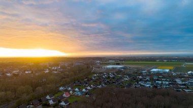 Ufukta parlayan turuncu ışık, aşağıda dağınık evler, uzaktaki yeşil alanlar ve gökyüzünün büyük kısmını kaplayan dramatik bulutlar ile gün doğumunda sakin bir kasabanın hava görüntüsü..