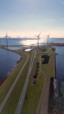 Yeşil tarlalarda rüzgar türbinleriyle kaplı iki su kütlesi arasında bir kıyı yolu var. Sahnede yenilenebilir enerji, altyapı ve doğa açık mavi gökyüzü altında vurgulanıyor..