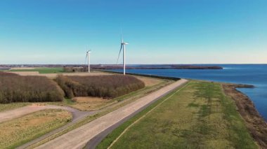 Yeşil tarlalarda rüzgar türbinleriyle kaplı iki su kütlesi arasında bir kıyı yolu var. Sahnede yenilenebilir enerji, altyapı ve doğa açık mavi gökyüzü altında vurgulanıyor..