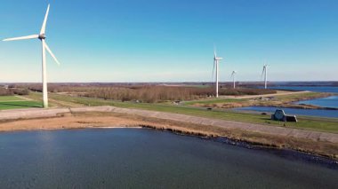 Yeşil tarlalarda rüzgar türbinleriyle kaplı iki su kütlesi arasında bir kıyı yolu var. Sahnede yenilenebilir enerji, altyapı ve doğa açık mavi gökyüzü altında vurgulanıyor..
