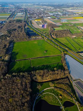 Yeşil tarlaları, ormanları, göletleri ve uzak bir kasabası olan kırsal bir arazinin havadan görünüşü. Renkli çiçek tarlaları ve açık mavi gökyüzünün altında uzanan tarlalar..