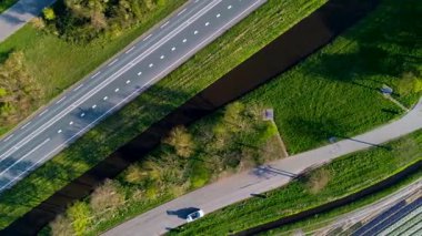 Rengarenk bir çiçek tarlasının yukarıdan aşağı manzarası düzgün sıralanmış sıralar, kıvrımlı bir kırsal yol, bir kanal ve uzun sabah gölgeleri olan hareket eden arabaların olduğu bir otoyol. Huzurlu bahar sahnesi.