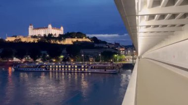 Bratislava Kalesi 'nin manzaralı bir gece manzarası, Tuna Nehri' ne bakan bir tepenin tepesinde aydınlandı. Şehir ışıklarının yansımaları, nehir kıyısı boyunca yanaşmış teknelerle suda parıldıyor..