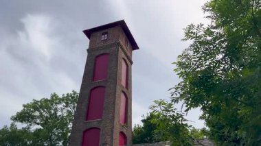 Kırmızı pencere kaplamalı uzun bir tuğla kule, yemyeşil ağaçlarla çevrili parçalı bulutlu bir gökyüzüne karşı duruyor. Sahne, mimariyle doğayı harmanlayan tarihi ve gizemli bir atmosfere sahiptir.