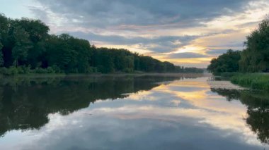 Sakin bir göl, yemyeşil ağaçlarla çevrili günbatımının altın tonlarını yansıtır. Suda hafif dalgalanmalar oluşur ve gökyüzü yumuşak maviler ve ılık portakalların karışımıdır. Huzurlu bir manzara yaratır.