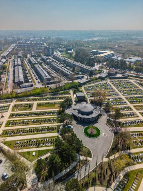 Yuvarlak yolları ve mezar taşlarıyla çevrili, ağaçlarla çevrili ve açık mavi gökyüzünün altındaki yerleşim alanına komşu büyük, düzenli bir mezarlığın havadan görünüşü..