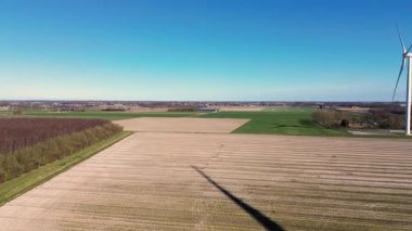 Kırsal bir alanda, yeşil tarlaları, bir nehri ve uzak bir köyü olan büyük bir rüzgar türbininin hava görüntüleri. Yel değirmeni açık mavi bir gökyüzü altında dönerek, doğadaki sürdürülebilir enerjiyi vurgular..