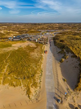 Sahil kenarındaki bir restoranın havadan görünüşü altın kum tepeleri ve yürüyüş yollarıyla çevrilidir. İnsanlar kıyıya yakın doğal bir yerde güneşli bir günün tadını çıkarırlar..