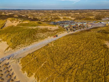 Sahil kenarındaki bir restoranın havadan görünüşü altın kum tepeleri ve yürüyüş yollarıyla çevrilidir. İnsanlar kıyıya yakın doğal bir yerde güneşli bir günün tadını çıkarırlar..
