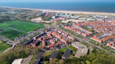 Kırmızı çatılı evleri, spor sahaları, yeşil alanları ve açık mavi gökyüzünün altındaki sahili gösteren bir sahil manzarası..