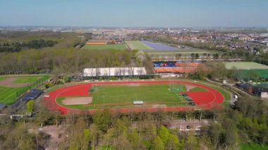 Açık mavi gökyüzünün altında yeşil tarlalar, spor kompleksleri, koşu yolları ve renkli tarım arazileriyle çevrili bir kasabanın havadan görünüşü, barışçıl ve organize edilmiş kırsal bir manzarayı gösteriyor..