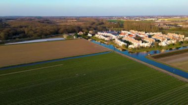 Etrafı tarlalar ve kanallarla çevrili huzurlu bir banliyö manzarası. Kırmızı çatıları olan düzenli evler suları yansıtıyor, tarlaların arka planına ve açık gökyüzüne karşı kuruluyor..
