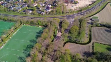 Ağaçlarla çevrili spor sahalarının, konut evlerinin, yolların ve tarım arazilerinin havadan görünüşü, açık gökyüzü altında kentsel ve kırsal manzaraların bir karışımını gösteriyor.