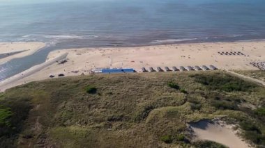 Sahil kulübeleriyle dolu güzel bir kumsal manzarası, kıyıda dinlenen insanlar ve ön planda çimenli kum tepeleriyle çevrili, kıyıya vuran nazik dalgalar..