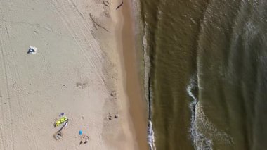 Yumuşak dalgaların kumlu sahille buluştuğu sahilin havadan görünüşü. İnsanlar kuma dağılmış durumda, bazıları kumdan kaleler yapıyor, diğerleri güneşli günün tadını çıkarıyor..