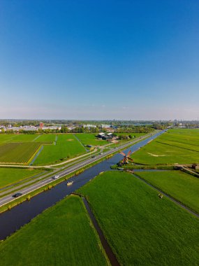 Geleneksel bir yel değirmeni olan Hollanda kırsalının havadan görünüşü. Kanal, yeşil tarlalar ve açık mavi gökyüzü altında hareket eden arabaların olduğu yakın bir yol..