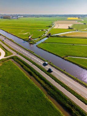 Geleneksel bir yel değirmeni olan Hollanda kırsalının havadan görünüşü. Kanal, yeşil tarlalar ve açık mavi gökyüzü altında hareket eden arabaların olduğu yakın bir yol..