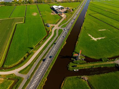 Geleneksel bir yel değirmeni olan Hollanda kırsalının havadan görünüşü. Kanal, yeşil tarlalar ve açık mavi gökyüzü altında hareket eden arabaların olduğu yakın bir yol..