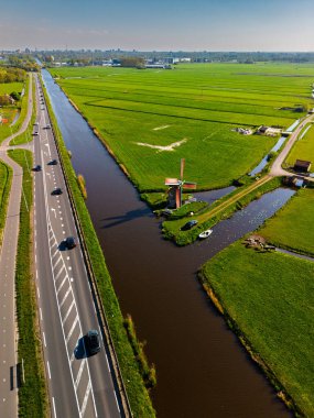 Geleneksel bir yel değirmeni olan Hollanda kırsalının havadan görünüşü. Kanal, yeşil tarlalar ve açık mavi gökyüzü altında hareket eden arabaların olduğu yakın bir yol..